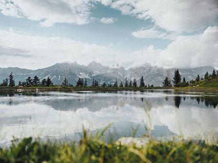 Entdecken Sie die Region mit der Wilder Kaiser GästeCard! Bergsee mit spiegelnder Wasseroberfläche und bewölktem Himmel