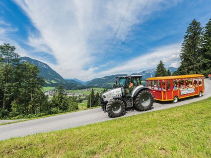 Entdecken Sie die Region mit der Wilder Kaiser GästeCard! Traktor zieht einen roten Touristenwagen mit Passagieren auf bergiger Straße