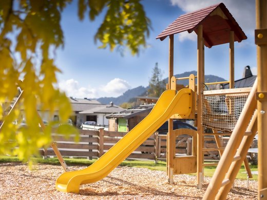 Kinderspielplatz mit Rutsche im Freien Ein sonniger Spielplatz im Freien mit einer gelben Rutsche, umgeben von Holzzäunen und malerischer Berglandschaft im Hintergrund.
