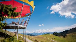 Sommer am Wilden Kaiser: Wandern und mehr Menschen auf Aussichtsplattform mit rotem Sonnenschutz und Berglandschaft im Hintergrund