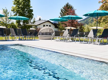Der Postwirt: Hotel mit Infinitypool in Tirol Pool mit Liegestühlen und blauen Sonnenschirmen in einem Garten bei Sonnenschein