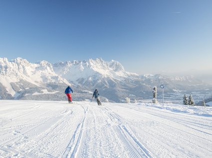 Winter in Söll: das Skigebiet SkiWelt Wilder Kaiser Zwei Skifahrer auf schneebedeckter Piste mit Alpen im Hintergrund bei klarem Himmel