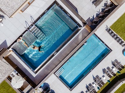 Zwei Außenpools im Hotel aus der Vogelperspektive Vogelperspektive auf zwei Außenpools in einem Hotel, einer mit Gästen im Wasser, umgeben von Liegestühlen und Sonnenschirmen auf einer modernen Poolterrasse.