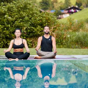 Urlaub in Söll mit Kindern oder als Pärchen Paar meditiert im Lotussitz vor einem Pool in grüner Landschaft