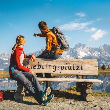 Urlaub in Söll mit Kindern oder als Pärchen Zwei Wanderer auf Bank vor Bergsee und Alpen mit Aufschrift Lieblingsplatzl