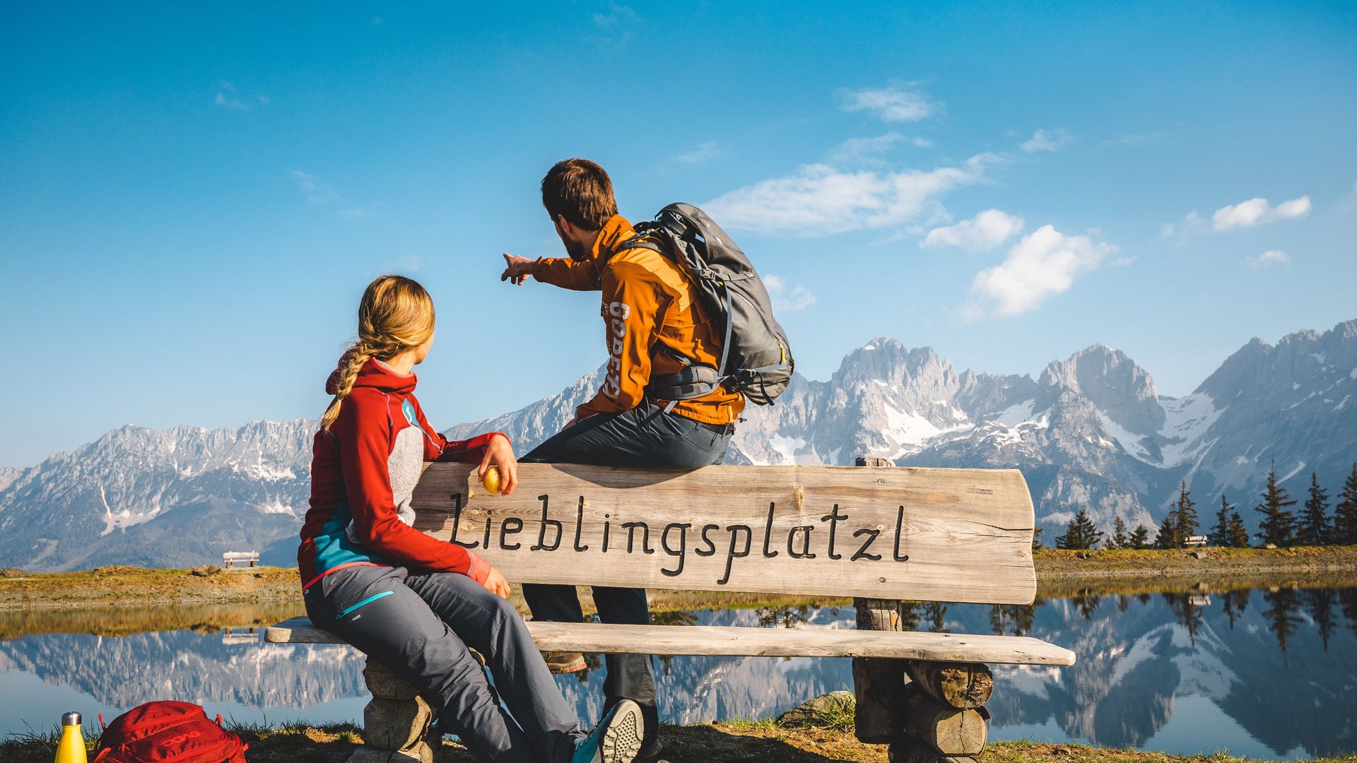 Entdecken Sie die Region mit der Wilder Kaiser GästeCard! Zwei Wanderer auf Bank vor Bergsee und Alpen mit Aufschrift Lieblingsplatzl