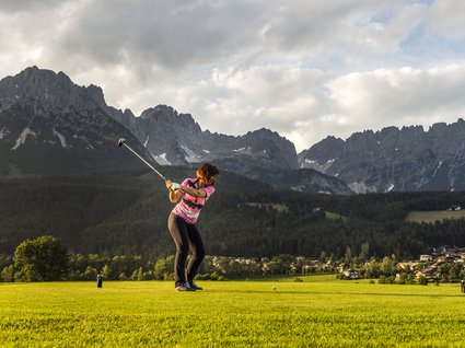Entdecken Sie die Region mit der Wilder Kaiser GästeCard! Frau spielt Golf vor einer Berglandschaft mit bewölktem Himmel