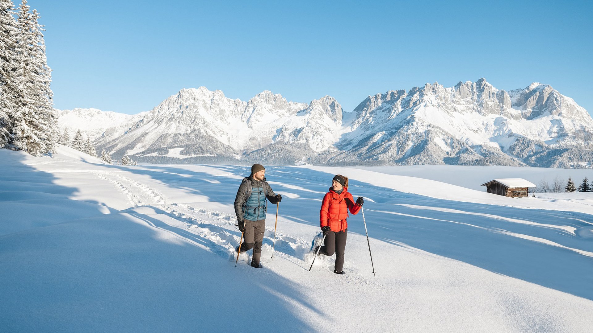 Entdecken Sie die Region mit der Wilder Kaiser GästeCard! Zwei Personen bei Winterwanderung im Schnee vor verschneiten Bergen und blauem Himmel