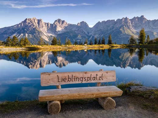 Entdecken Sie die Region mit der Wilder Kaiser GästeCard! Bank mit Aufschrift Lieblingsplatz vor Bergsee mit Spiegelung der Berge