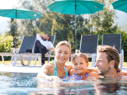 Der Postwirt: Hotel mit Infinitypool in Tirol Familie mit Kind entspannt im Pool neben Liegestühlen und Sonnenschirmen