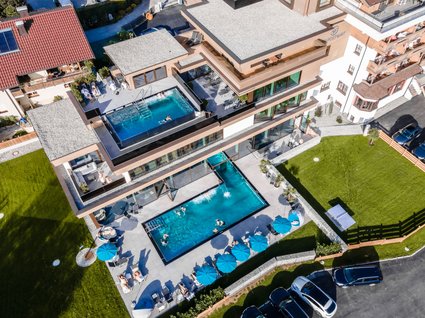 Ihre Traumtage im Postwirt Söll Luftaufnahme eines Hotels mit zwei Außenpools und Liegestühlen mit blauen Sonnenschirmen