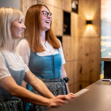 Urlaub in Söll mit Kindern oder als Pärchen Zwei Frauen in Trachten lächeln an einer Rezeption in einem Holzinterieur