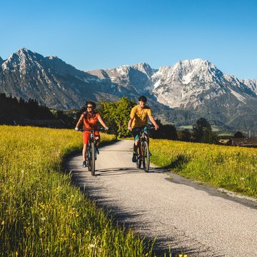 Urlaub in Söll mit Kindern oder als Pärchen Zwei Radfahrer auf Landstraße vor Berglandschaft mit grünen Wiesen und blühenden Blumen