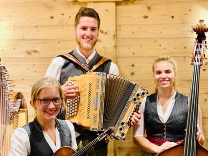 Kulinarische Verführungen in unserem Hotel in Söll/Tirol Drei Musiker in Trachten mit Geige, Akkordeon und Kontrabass vor Holzwand