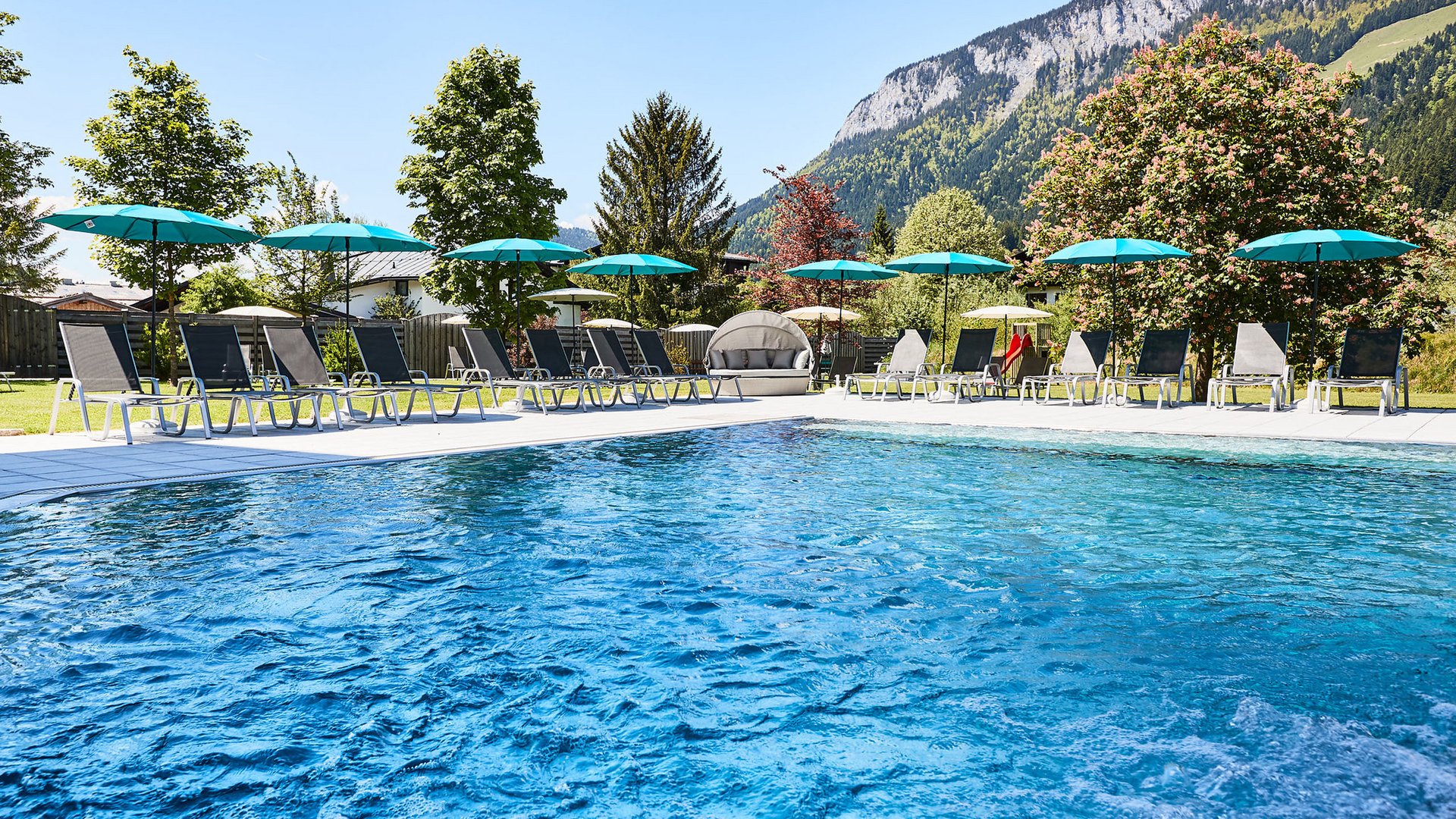 Der Postwirt: Hotel mit Infinitypool in Tirol Außenpool mit Liegestühlen und Sonnenschirmen vor Berglandschaft an sonnigem Tag