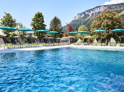 Ihre Traumtage im Postwirt Söll Außenpool mit Liegestühlen und Sonnenschirmen vor Berglandschaft an sonnigem Tag