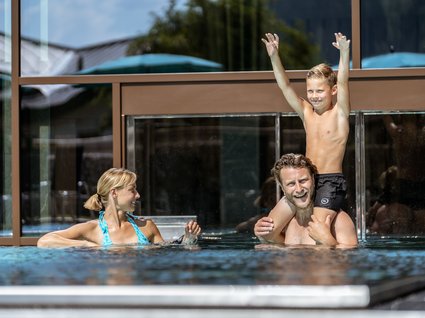 Familie spielt im Hotelpool Ein Junge sitzt auf den Schultern seines Vaters, während die Mutter lächelnd im Pool schwimmt. Die Familie genießt die Zeit zusammen im Außenbereich eines Hotelpools.