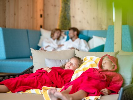 Kinder im Ruhebereich des Spas Zwei Kinder in roten Bademänteln schlafen entspannt im Spa-Ruhebereich, während ihre Eltern im Hintergrund auf einer Couch sitzen.