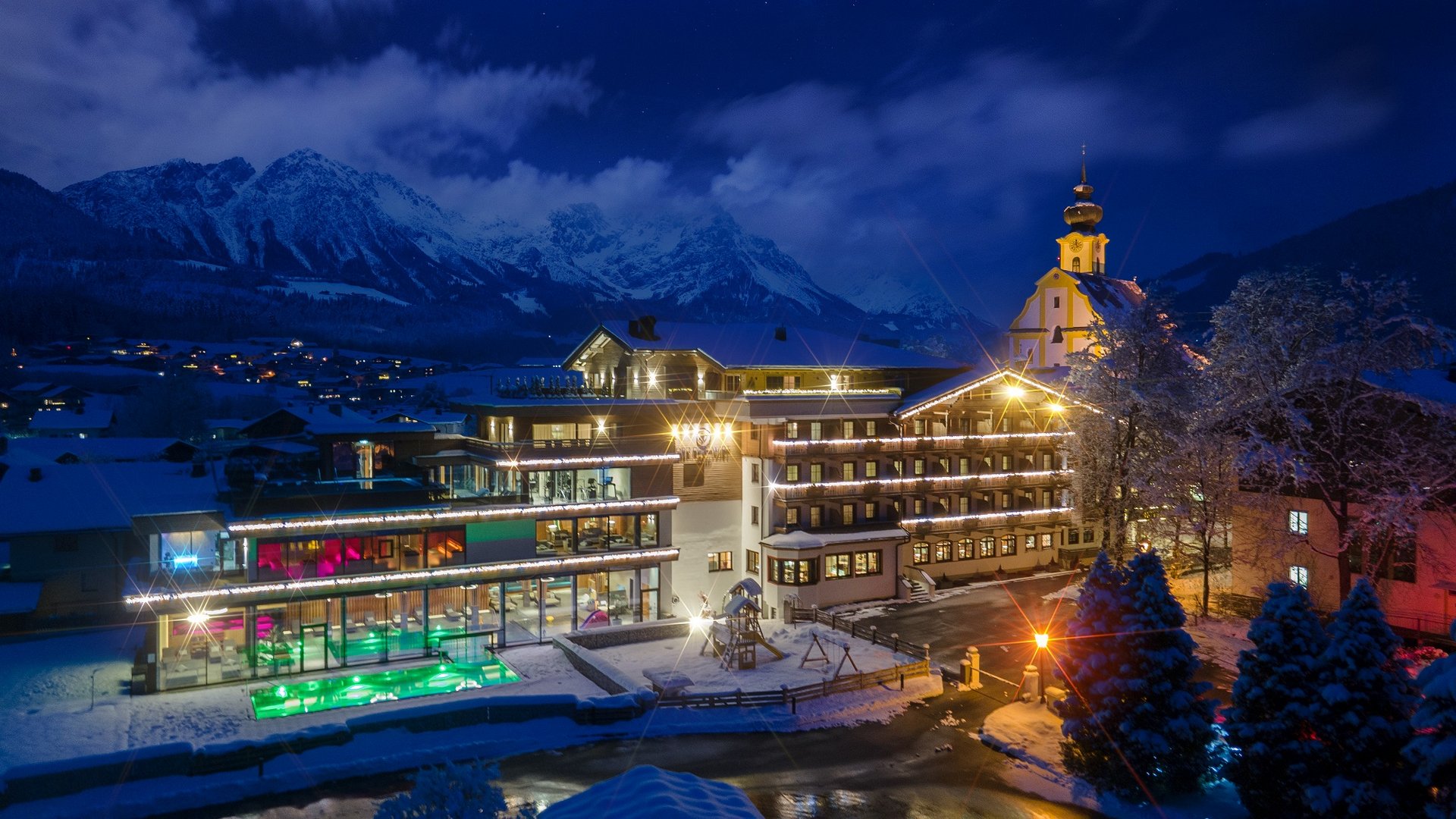 Hotel in Söll am Wilden Kaiser: der Postwirt Beleuchtetes Hotel und Kirche in verschneitem Tal vor Bergpanorama bei Nacht