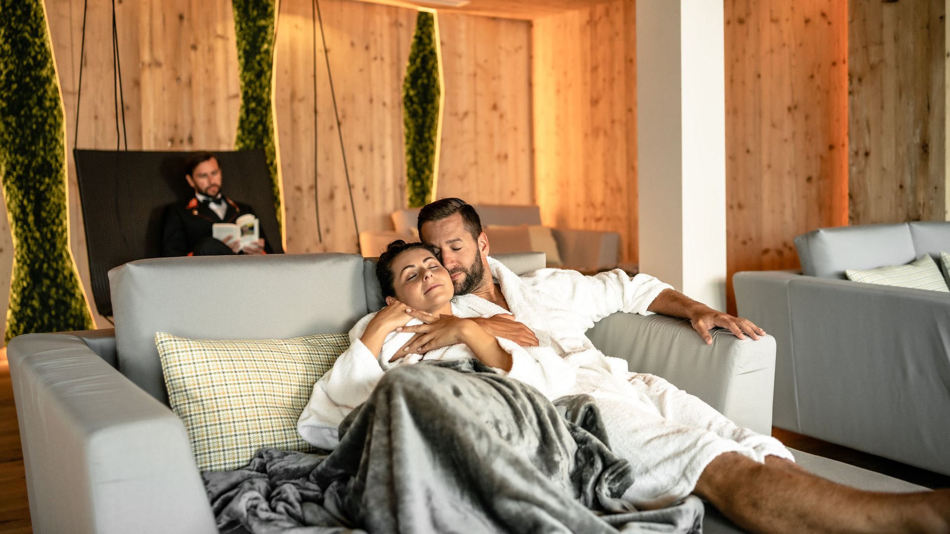 Wellnesshotel in Söll Paar in Bademänteln entspannt auf Sofa in gemütlichem Spa mit Holzinterieur