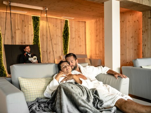 Ihre Traumtage im Postwirt Söll Paar in Bademänteln entspannt auf Sofa in gemütlichem Spa mit Holzinterieur