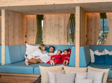 Ihre Traumtage im Postwirt Söll Familie entspannt in Bademänteln auf blauem Sofa in modernem Holzzimmer