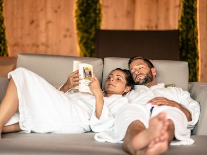 Entspannung im Wellnessbereich für Paare Paar in Bademänteln entspannt auf Sofa, Frau liest Buch, Mann schläft