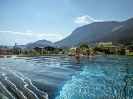 Infinity-Pool mit Alpenblick Gäste schwimmen im Infinity-Pool eines Hotels mit atemberaubendem Blick auf die umliegenden Berge und Täler bei strahlendem Sonnenschein.