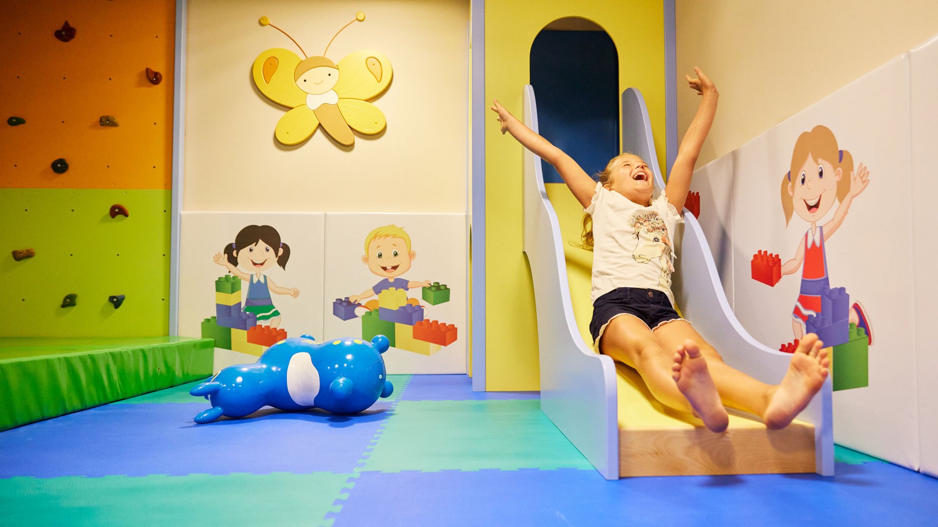Spaß und Action im Kinderhotel in Söll Fröhliches Mädchen rutscht in buntem Indoor-Spielbereich mit Wandbildern und Spielzeug