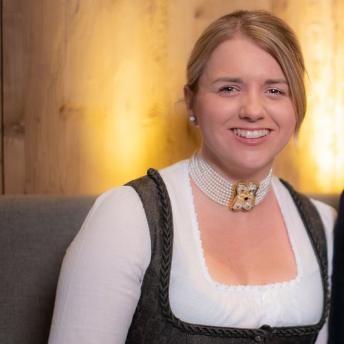 Unser Hotel in Söll/Österreich: eine spannende Geschichte Porträt eines lächelnden Paares in traditioneller bayerischer Tracht vor Holzwand