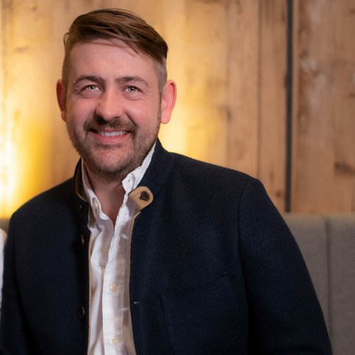 Unser Hotel in Söll/Österreich: eine spannende Geschichte Porträt eines lächelnden Paares in traditioneller bayerischer Tracht vor Holzwand