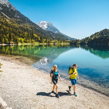 Urlaub in Söll mit Kindern oder als Pärchen Paar wandert am Ufer eines alpinen Sees mit Bergen und blauem Himmel im Hintergrund