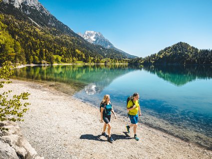 Entdecken Sie die Region mit der Wilder Kaiser GästeCard! Paar wandert am Ufer eines alpinen Sees mit Bergen und blauem Himmel im Hintergrund