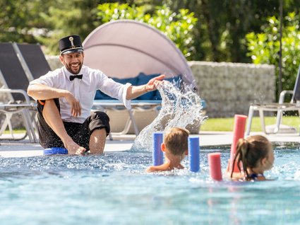 Kinder spielen mit Betreuer im Pool Kinder haben Spaß im flachen Pool, während ein Betreuer in einem lustigen Outfit mit ihnen spielt und Wasser spritzt, umgeben von Liegestühlen im Außenbereich.